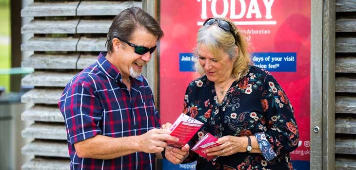 Two people explore the benefits of becoming a member by reading a membership leaflet while standing outside the Welcome Building at Westonbirt Arboretum