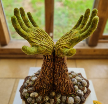 ‘Helping Hands’ is a life-sized felted sculpture of forearms and hands. The arms are upright, in the style of a tree trunk, with the hands opening out at the top, like branches of a tree. At the base of the tree are felted pebbles in shades of grey, some are textured with open weave cotton fabric over the surface. The trunk of the tree (arms) is covered in brown shades of felt, highly textured, like bark. Some of this extends into the pebbles, as roots. At the wrists, the colour of the felt changes to shades of green. The same bark-like texture extends over the palms and backs of the hands. This piece was inspired by a Blue Atlas Cedar within the Arboretum. The shape of the branches reminded artist Eleanor May of arms reaching out, in a gesture of both offering and requesting help. This led her to think of the symbiotic relationship between humans and Nature. The piece is highly textured and invites the viewer to explore the many varied textures to be found within the Arboretum. Credit: Eleanor May.