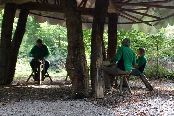 A group of volunteers crafting wood under a sheltered canopy using shaving horses.